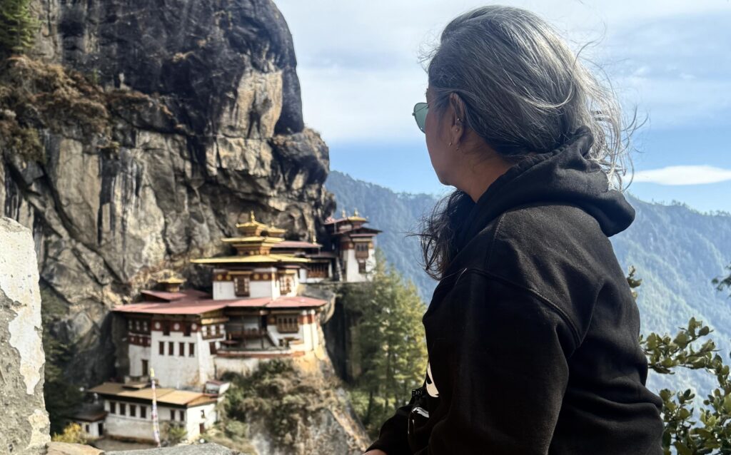 Madhurie Pandit, founder of Wheel to Heal, gazes toward the iconic Tiger’s Nest Monastery in Bhutan, nestled against a dramatic Himalayan cliff.